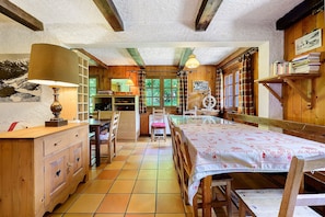 Habitació rústica amb taula de fusta, cadires, una panxa, làmpada, prestatge i finestra amb cortines.