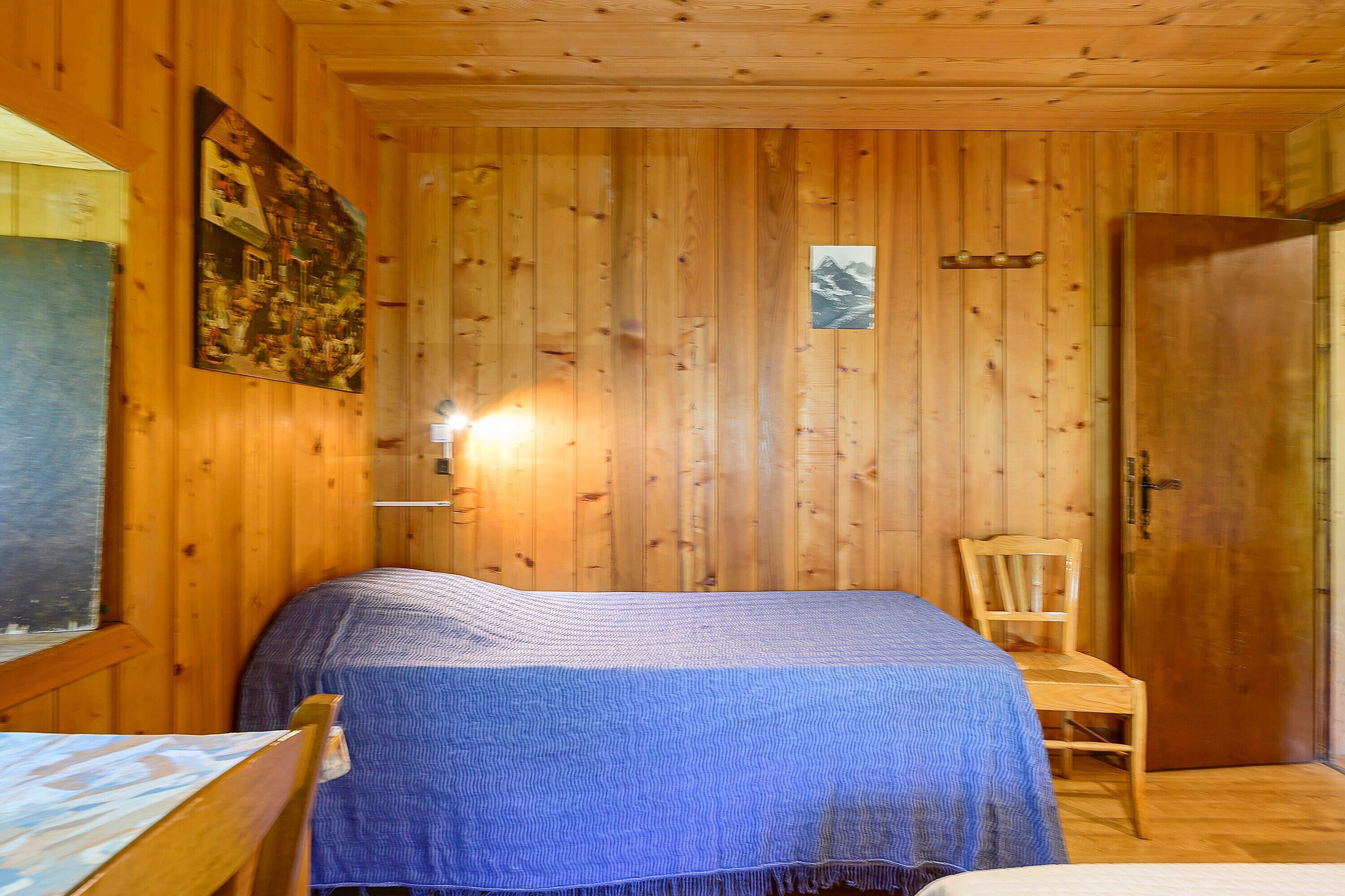 A wooden interior with a bed, a chair, a wall-mounted light, and a door.