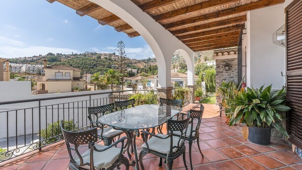 A terrace with a glass table and chairs, surrounded by potted plants and a view of a hillside town.