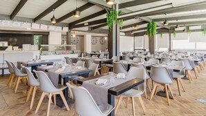 Un grand espace dédié à la salle à manger, avec plusieurs tables et chaises, une cuisine à l’arrière-plan et des plantes suspendues.