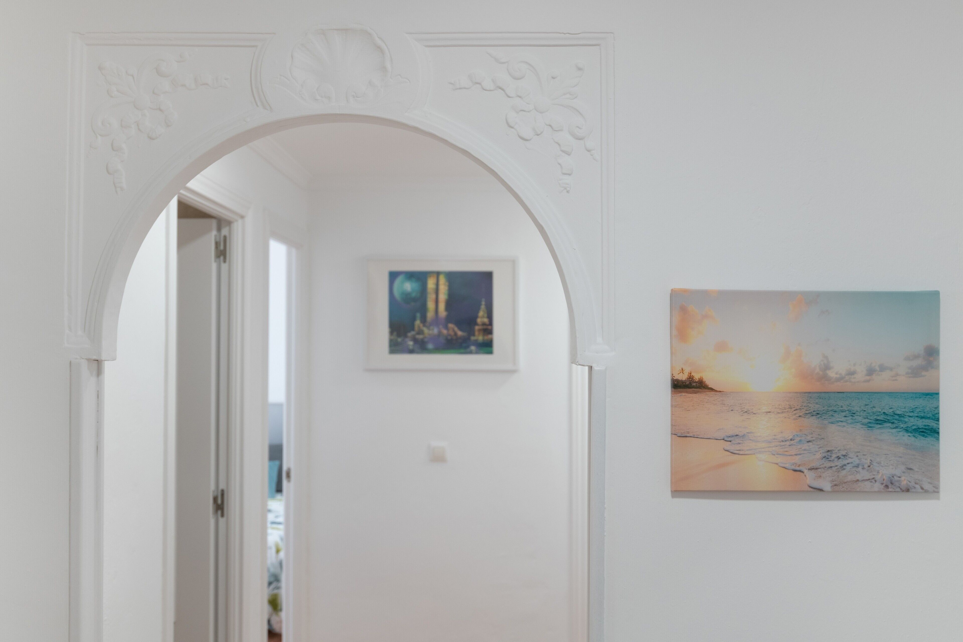 A hallway with an arched doorway, a framed painting of a sunset over the ocean, and a door leading to another room.