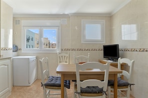 A kitchen with a dining table, chairs, a washing machine, a TV, and windows with a view of buildings.