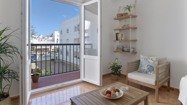 Un balcón con vistas a edificios, una mesa de madera con un plato decorativo, una silla de mimbre con cojín, y una estantería con libros y plantas.