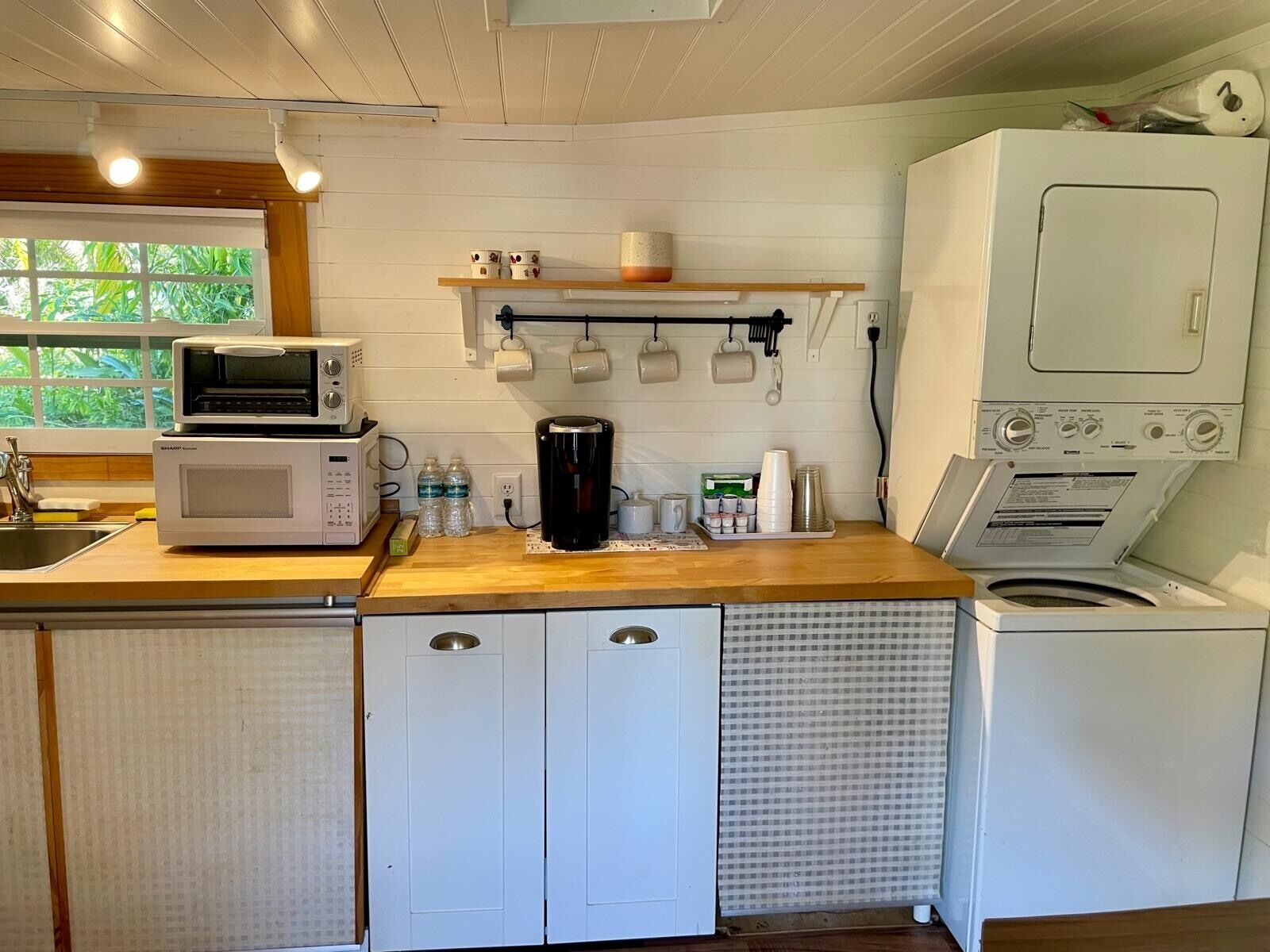 Cocina compacta equipada con microondas, cafetera y lavadora. Se observan armarios y una ventana con vegetación.
