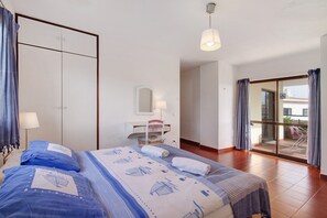 A bedroom with a bed, a desk, a chair, a mirror, and a sliding glass door leading to an outdoor area.