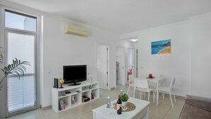 Un salon moderne avec un téléviseur à écran plat, une table basse sur laquelle se trouvent une bouteille et un verre, un coin repas avec des chaises blanches, et un tableau représentant une scène de plage.