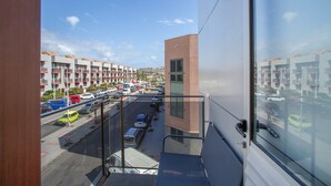Une vue depuis un balcon donnant sur un parking où sont stationnés de nombreux véhicules, sur un immeuble doté de balcons, et sur un ciel dégagé.