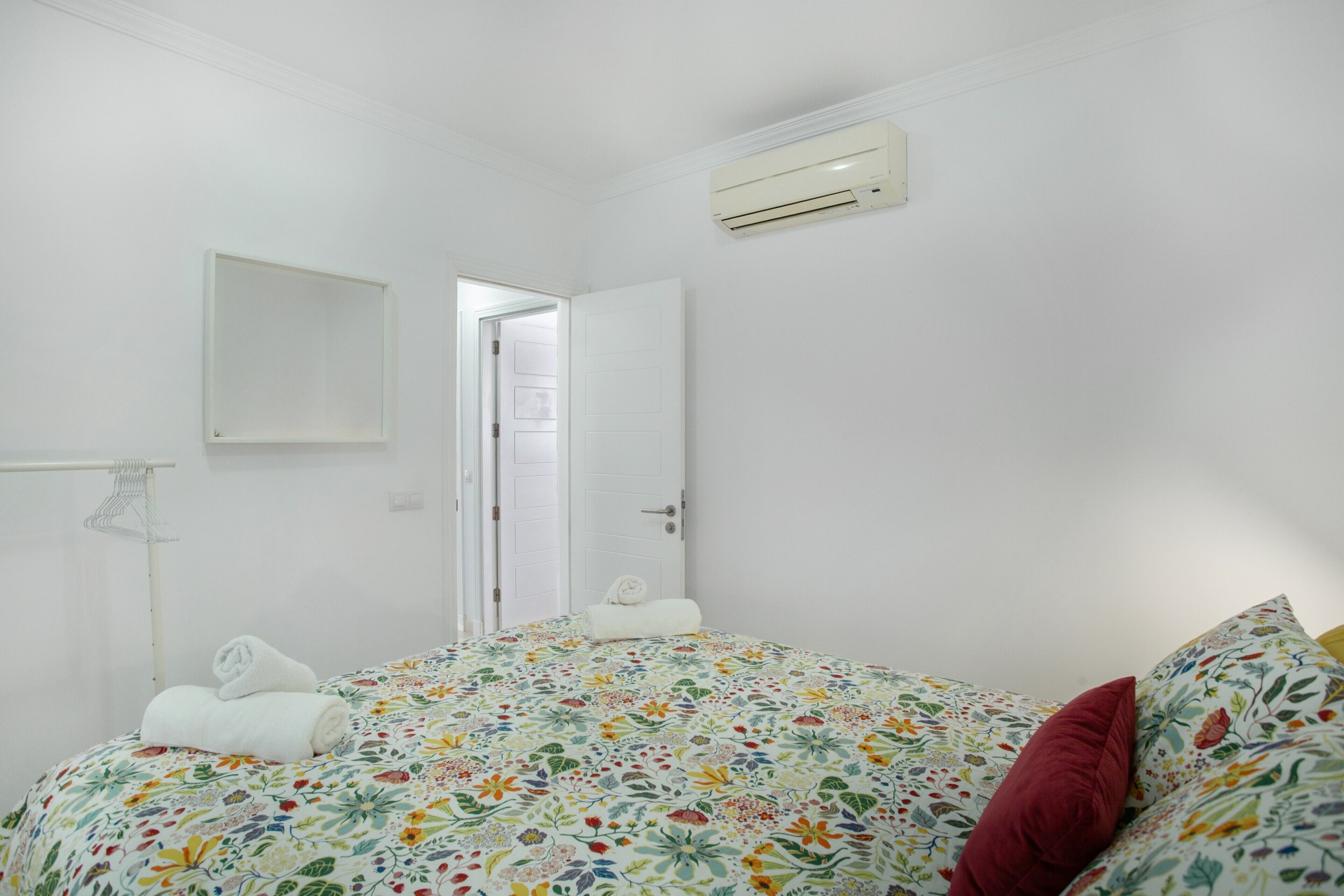 Un dormitorio con una colcha estampada con motivos florales, una almohada roja, una toalla blanca y un equipo de aire acondicionado blanco en la pared.
