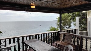 Ett takförsett utomhusområde med sittplatser och utsikt över havet, möbler i trä och ett räcke.