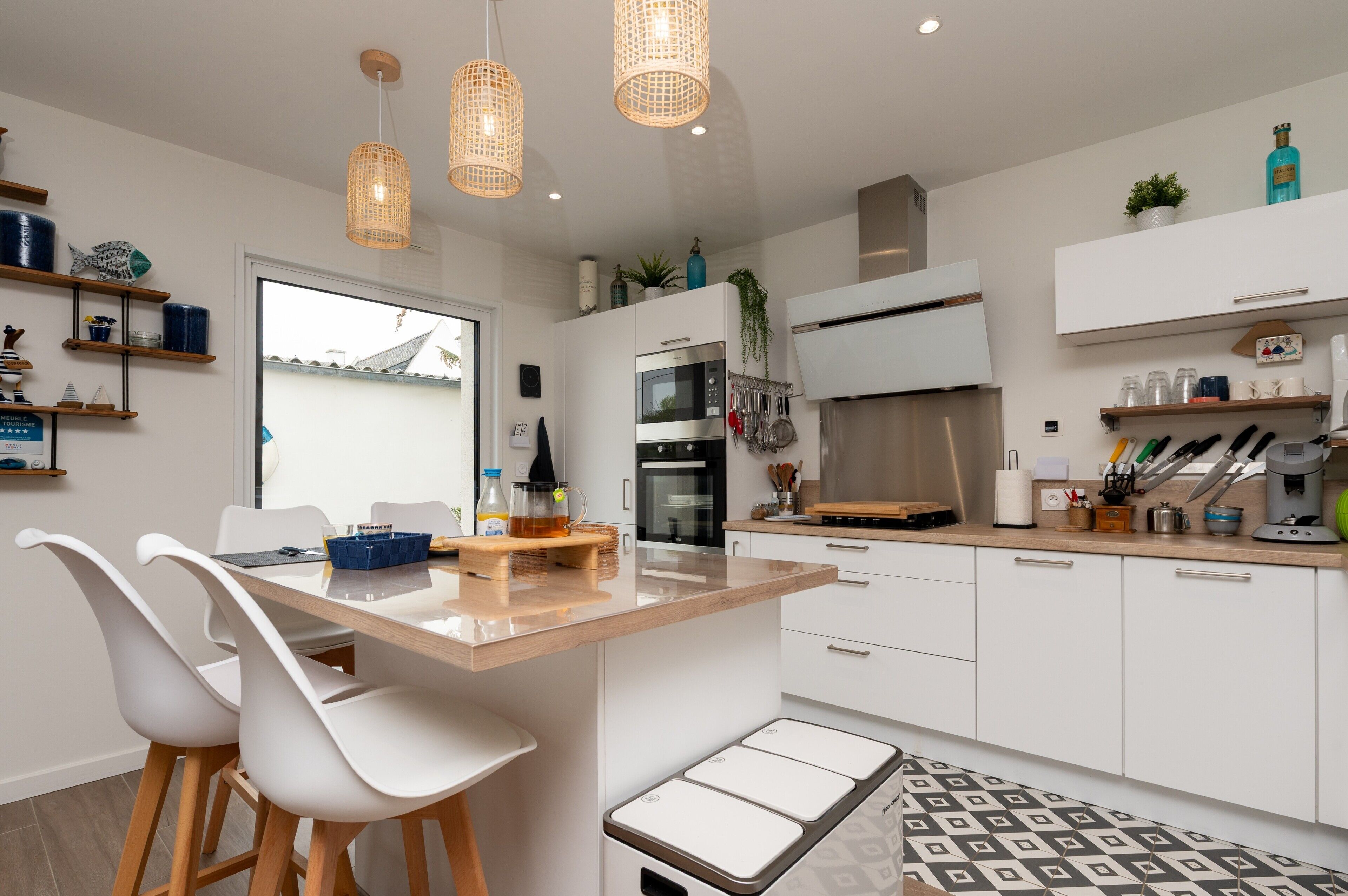 Une cuisine moderne avec des meubles blancs, un îlot central, des luminaires suspendus et une vue sur l’extérieur.