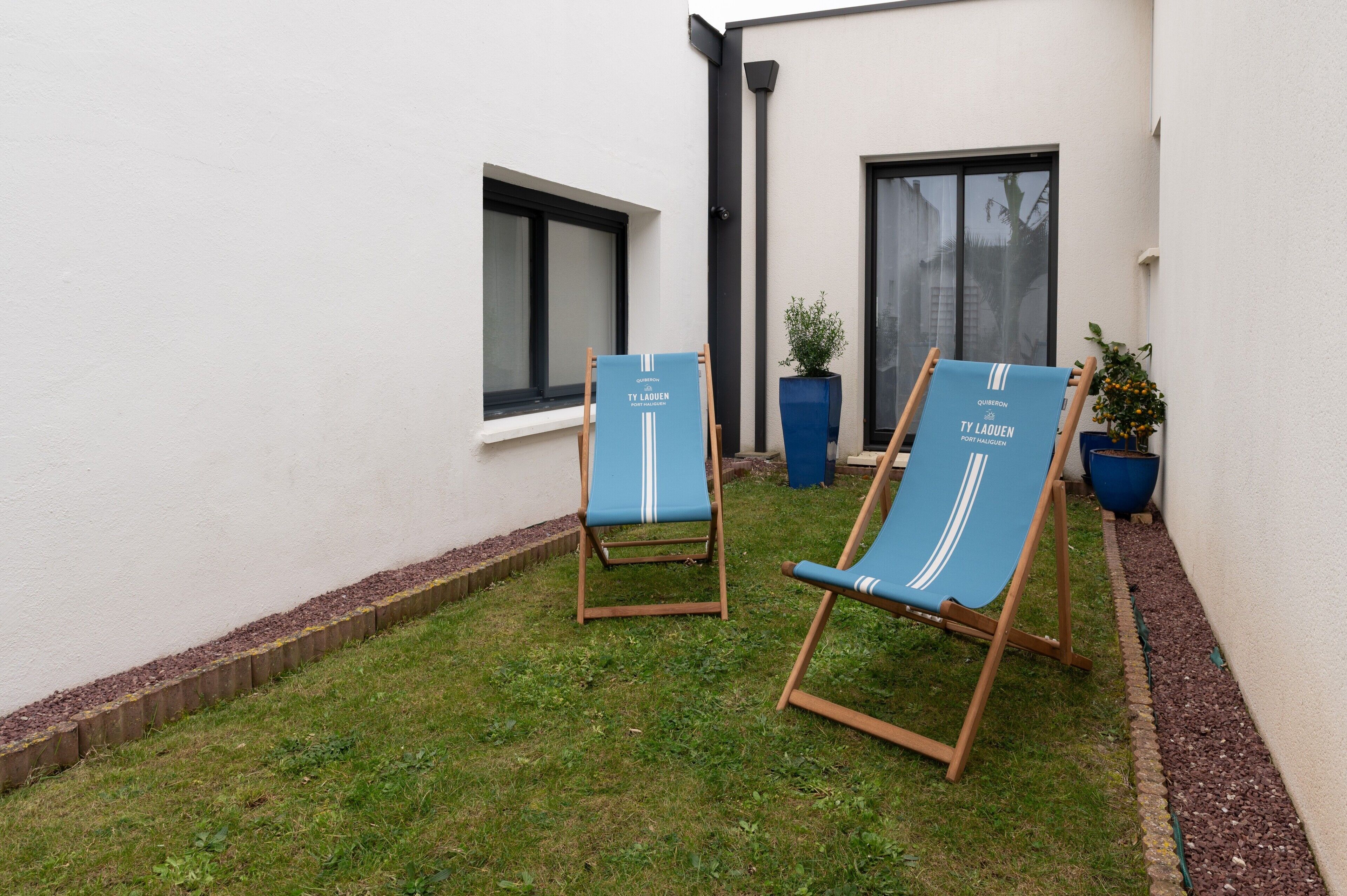 Deux fauteuils bleus confortables, portant la marque « TY LAUEN », sont placés dans un petit espace extérieur verdoyant, avec un mur blanc et des fenêtres.