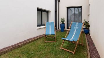 Deux fauteuils bleus confortables, portant la marque « TY LAUEN », sont placés dans un petit espace extérieur verdoyant, avec un mur blanc et des fenêtres.