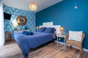 Un dormitorio con una pared azul estampada, una cama con ropa de cama azul, una silla de mimbre, un espejo redondo y un televisor en la pared.