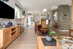 Una sala de estar moderna con televisor, muebles de madera, una zona de comedor con sillas y una cocina al fondo.