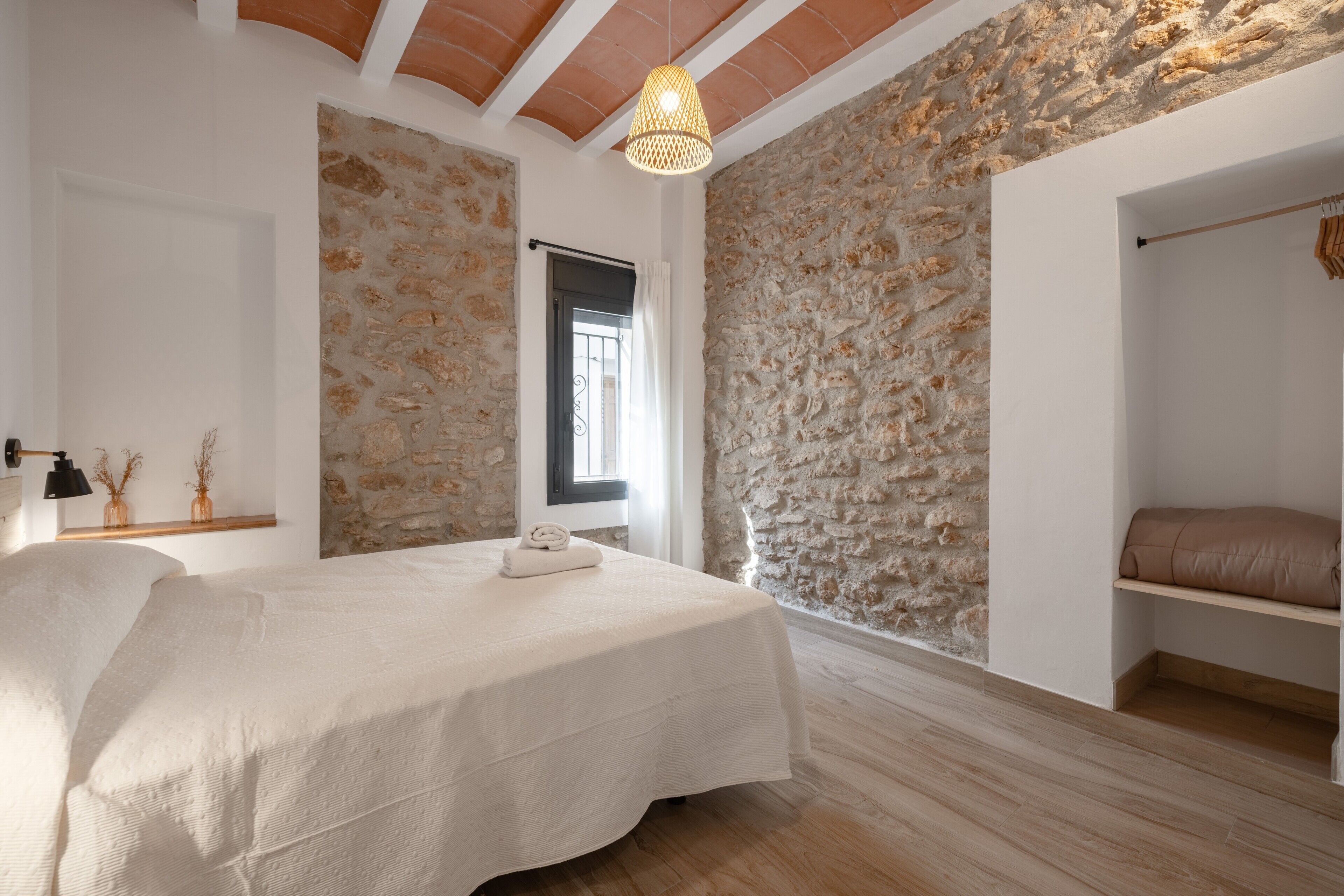 Une chambre avec un mur en pierre, un lit recouvert d’une courtepointe blanche, un luminaire suspendu et un plancher en bois.