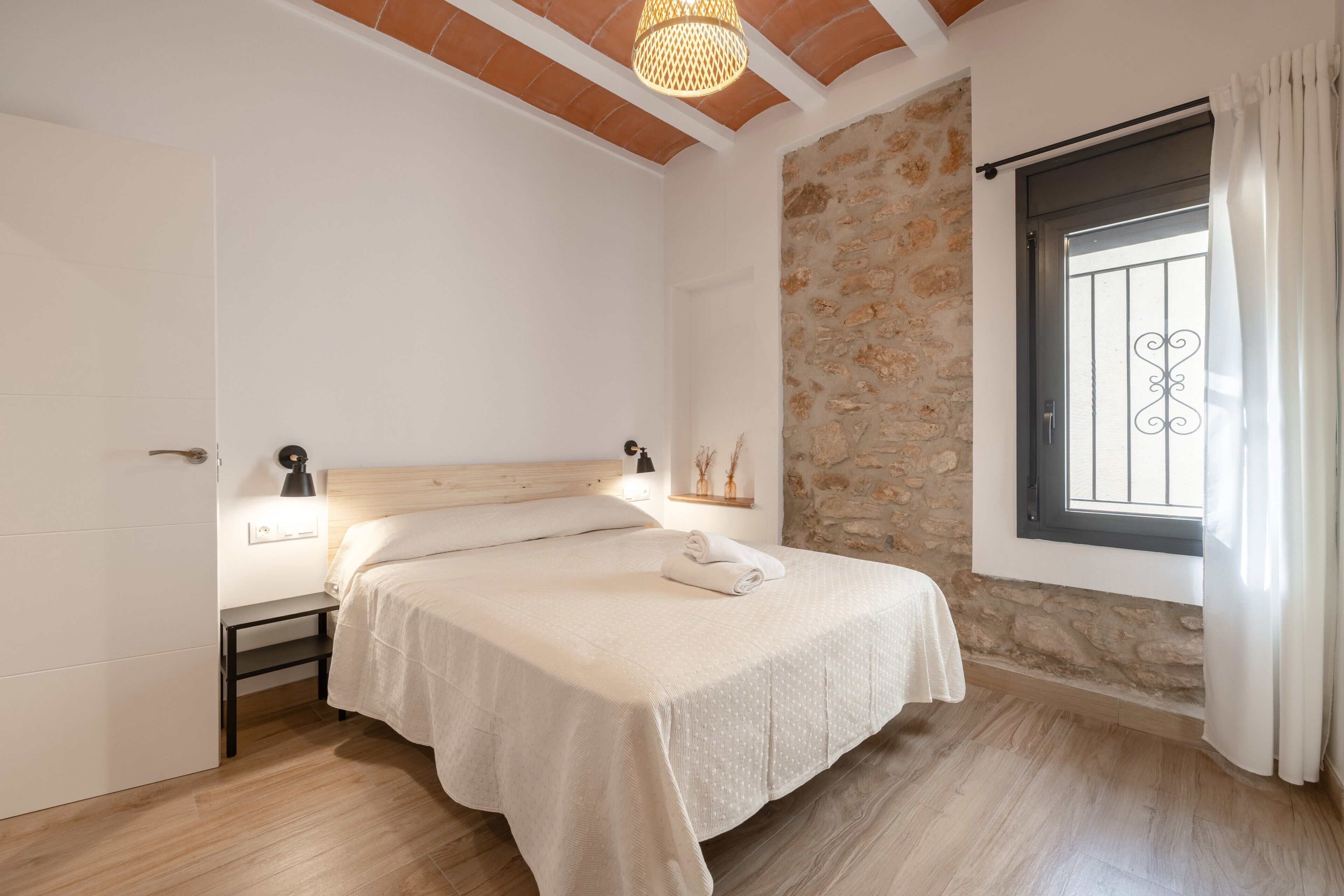 Un dormitorio con cama, mesitas de noche, pared de piedra, ventana con cortinas y techo de madera.