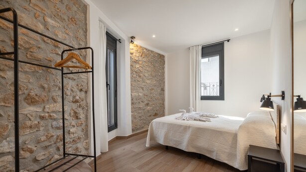 Een slaapkamer met een stenen muur, een bed met een witte dekbedovertrek, een houten kledingrek en een raam met gordijnen.