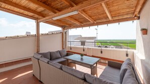 Een overdekt terras met een tafel, banken en uitzicht op een sportveld, afgedekt met een houten plafond.