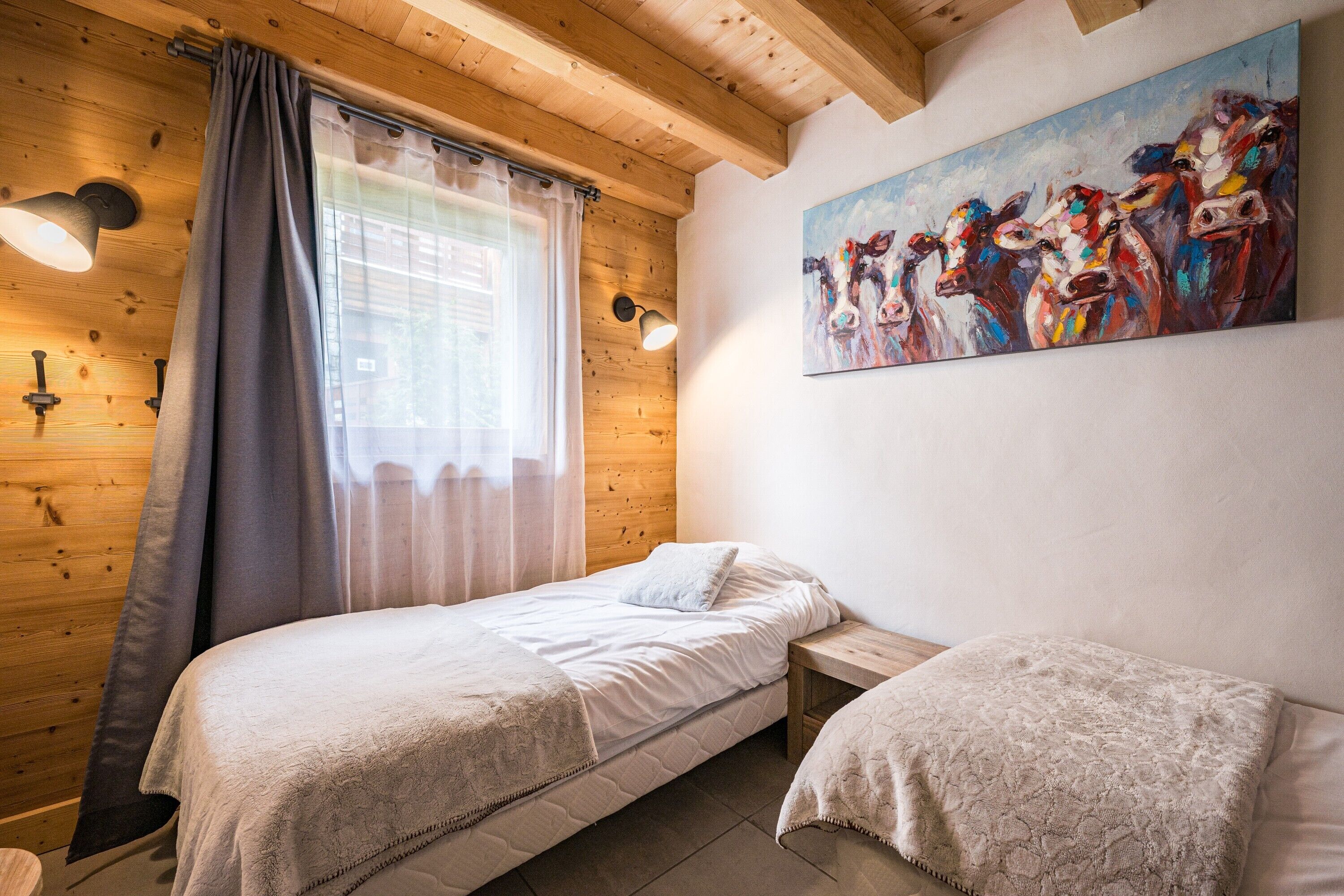 Une chambre avec un plafond en bois, un lit recouvert d’une couette blanche, une table de chevet, une commode, un tableau accroché au mur et une fenêtre avec des rideaux.