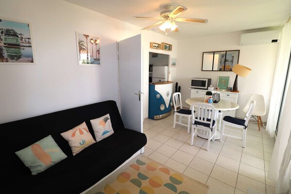 A compact living space with a black sofa, a dining area with a small table and chairs, a kitchenette, and a ceiling fan.