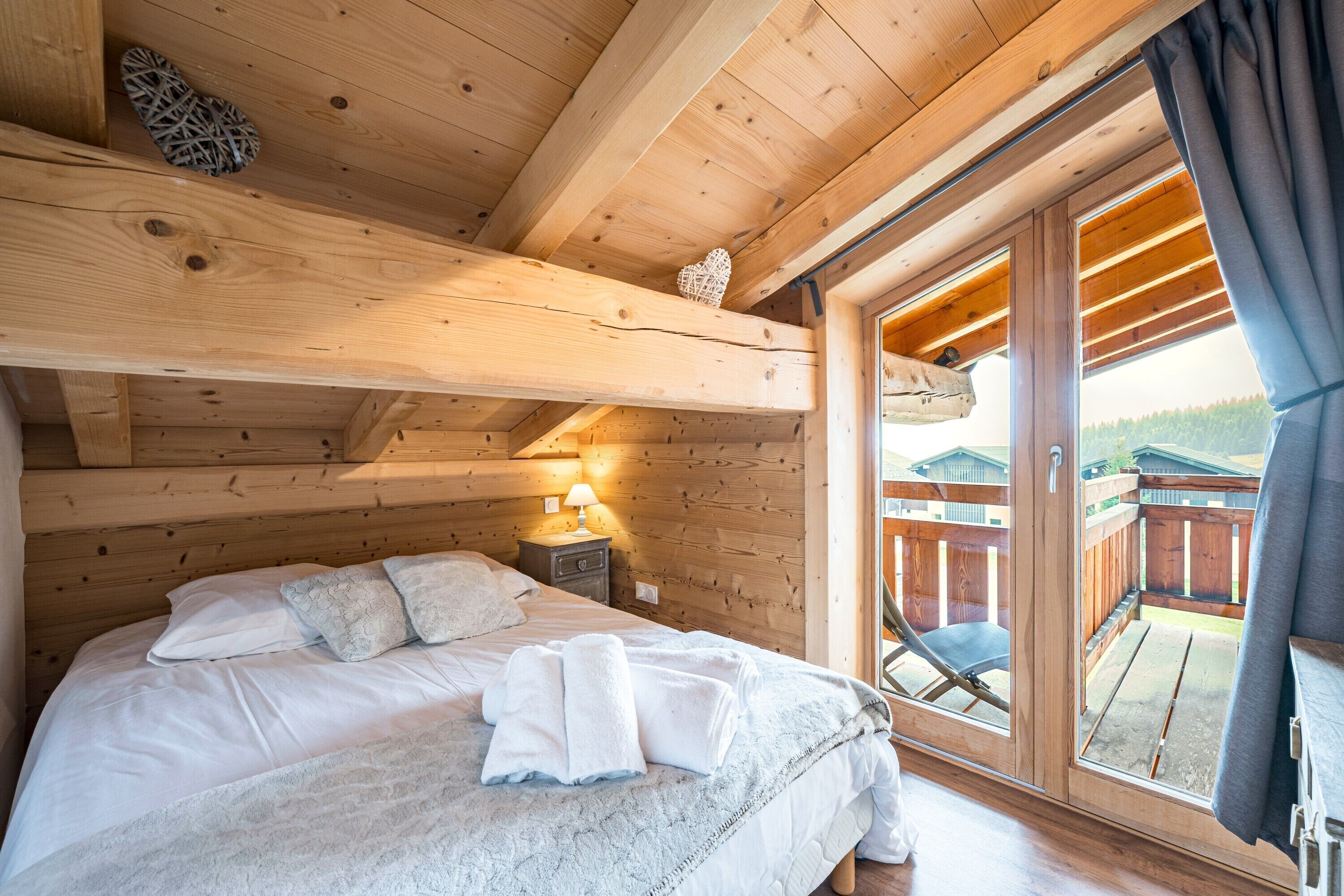 Un dormitorio de cabaña de madera con una cama, almohadas, una mesita de noche, una lámpara y una puerta corrediza de vidrio que da a una terraza exterior.