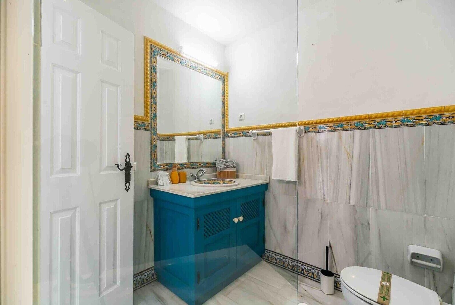 Casa de banho com porta branca, pia azul, espelho com moldura decorativa e cortina de chuveiro.