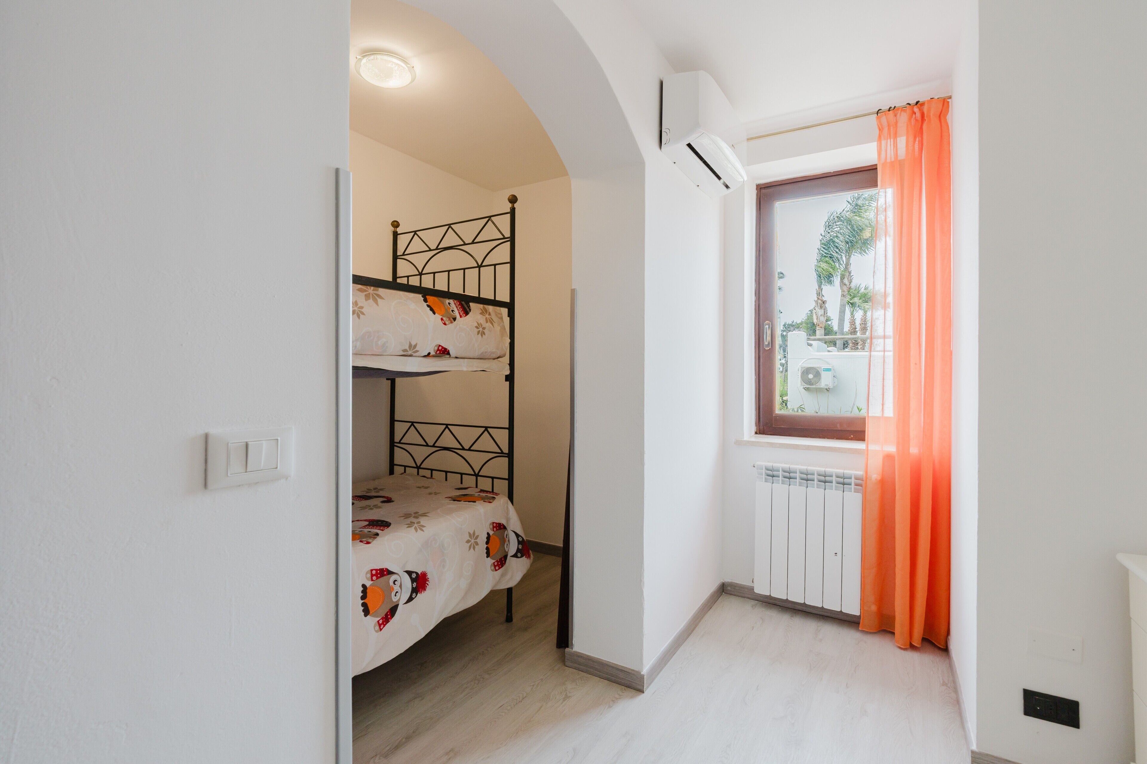Una habitación con literas, una ventana con cortinas, un radiador y un interruptor de luz en la pared.