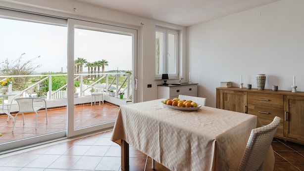 A balcony with a dining table, chairs, and a view of palm trees and the sea.
