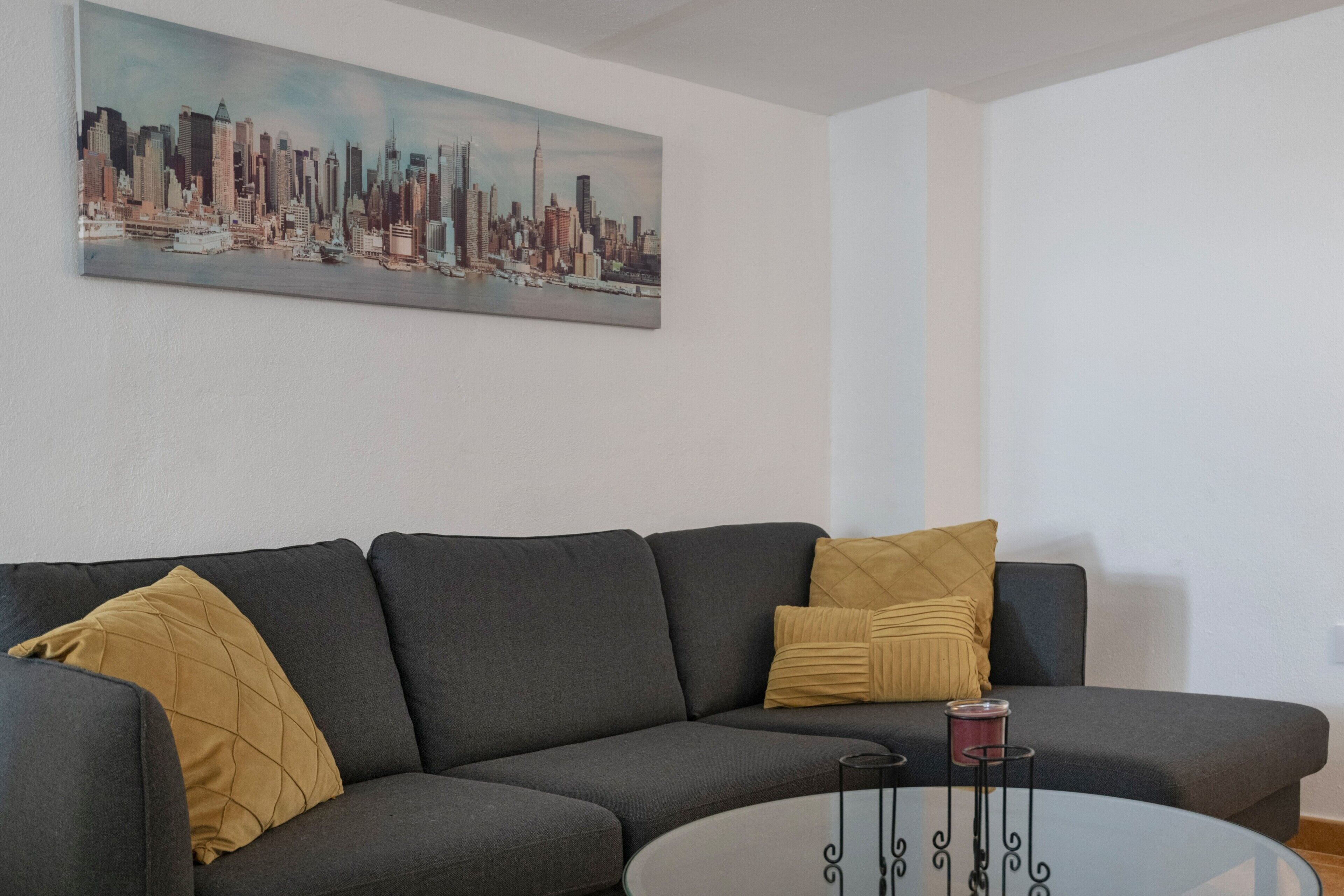 Una sala de estar con un sofá gris, dos cojines amarillos, una mesa de centro de vidrio y posavasos metálicos, y un cuadro de un paisaje urbano en la pared.
