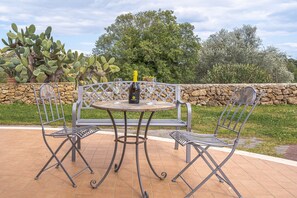 A patio with a round table and two chairs, a bottle of wine, and a stone wall.