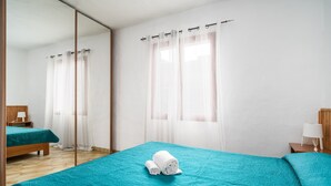 A hotel room with a bed, a mirror, a window with sheer curtains, and a wooden floor.