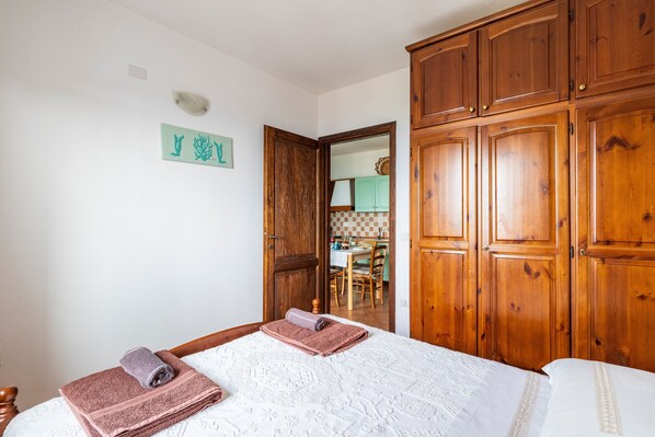 A bedroom with a bed, wooden wardrobe, and a view into the kitchen.