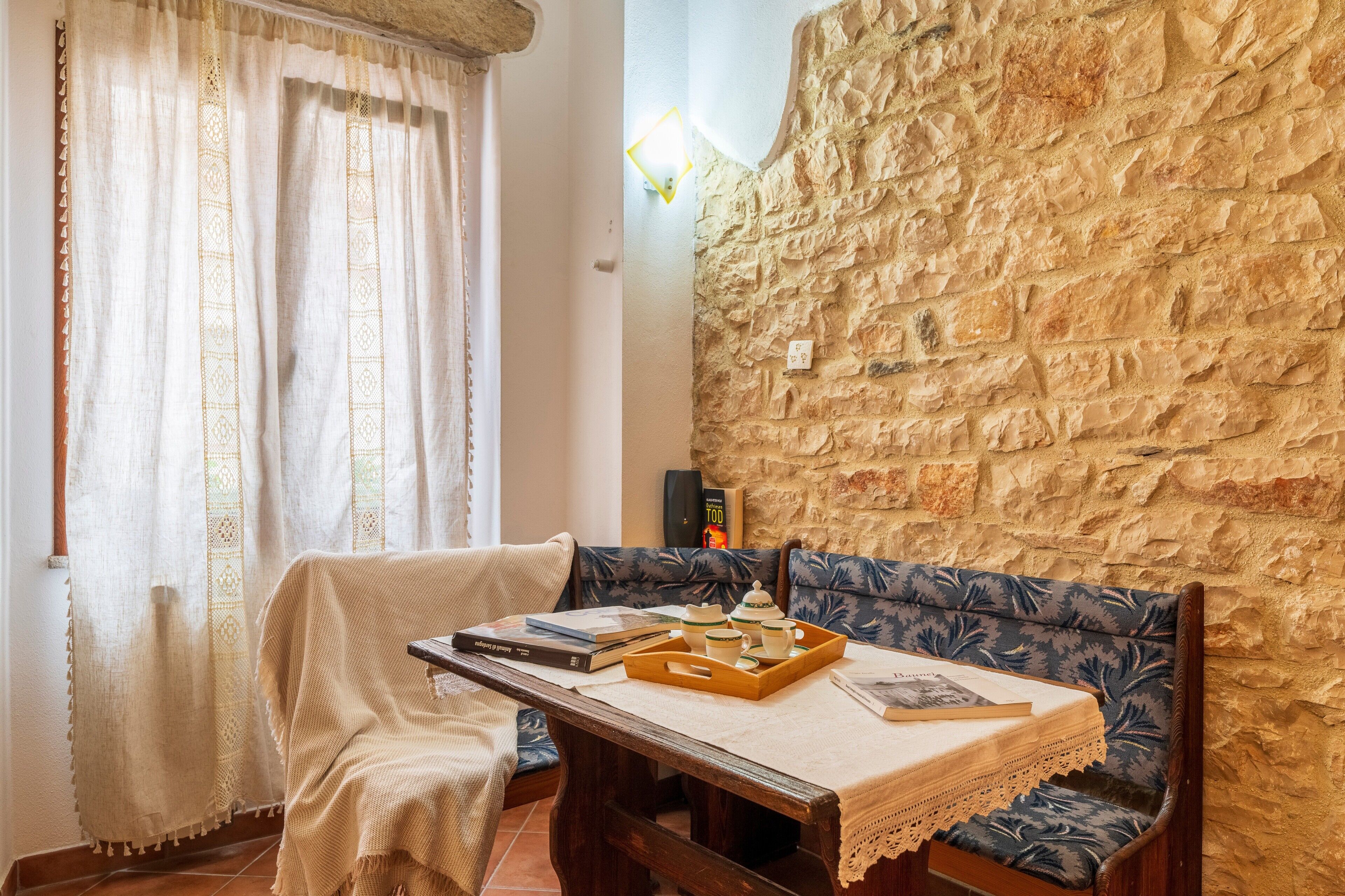 Una habitación acogedora con pared de piedra, un banco acolchado, una mesa de madera con una bandeja de tazas de té y una ventana con cortinas.