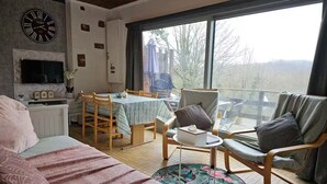 A cozy living room with a bed, a TV, a dining table, and a large window offering a view of trees. - Nawal's Belle Vue Dinant/Givet (Hastière)