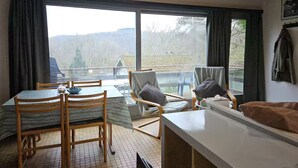 A room with a table, chairs, and a bed. There are large windows with curtains, and a view of the outdoors. - Nawal's Belle Vue Dinant/Givet (Hastière)