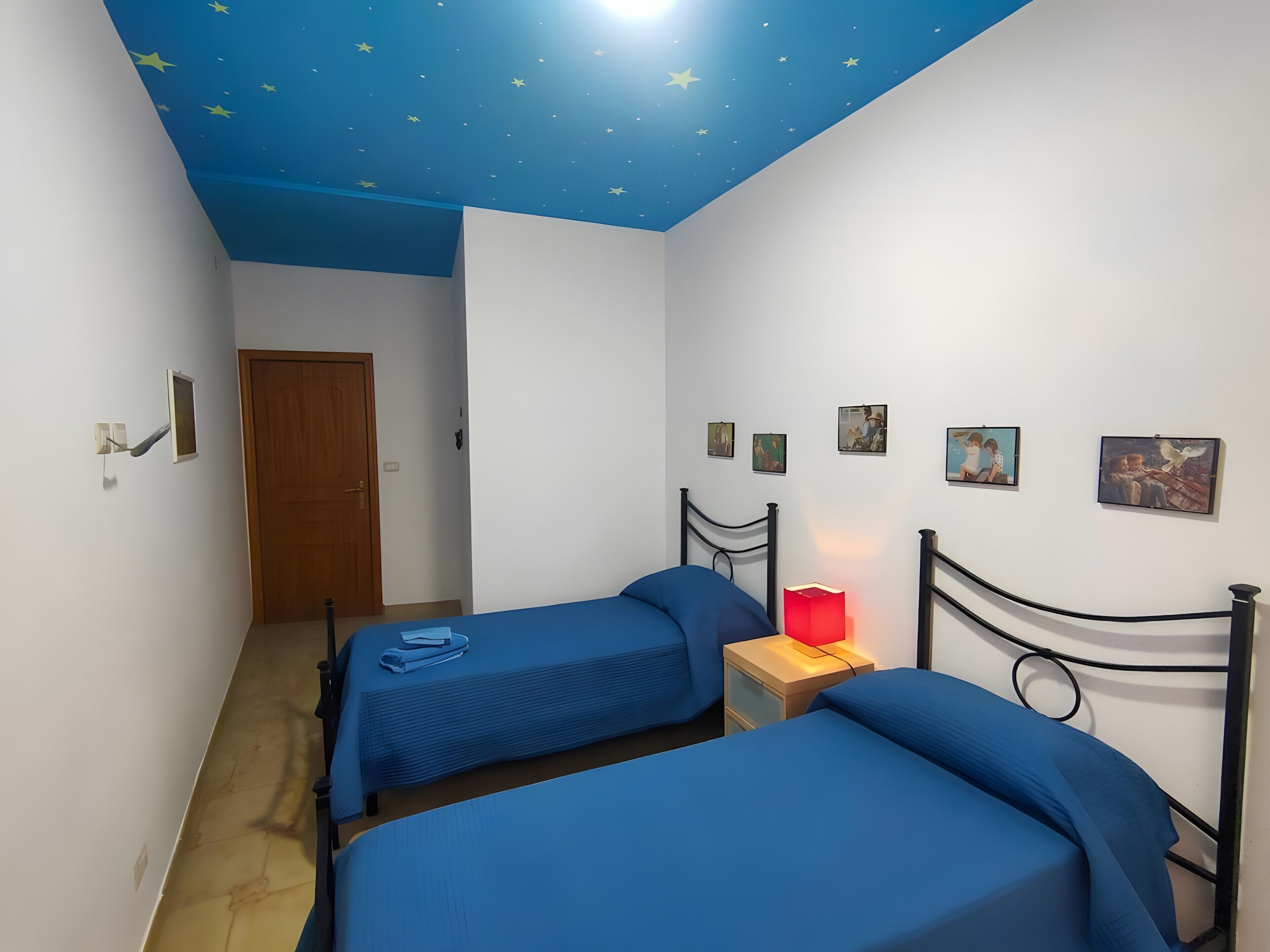 A room with two beds, a nightstand with a red lamp, and a door.