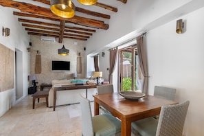 A modern interior with exposed wooden beams, a dining area with wicker chairs, a kitchen with a flat-screen TV, and a large window with curtains.