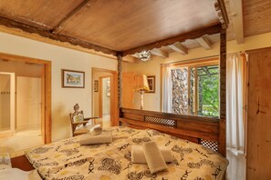 Una camera da letto con soffitto in legno, un letto con lenzuola decorate, un comodino, una lampada e una finestra vista su vegetazione.