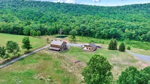 A rural property with two main buildings, a large barn-like structure and a smaller house, surrounded by dense forest and open grassy areas.