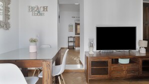 Una sala de estar moderna con una mesa de comedor de vidrio, sillas blancas, un televisor plano grande, mueble de entretenimiento de madera y un letrero en la pared que dice 'BEACH HOUSE'.