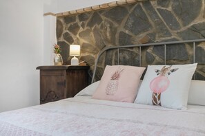 Una cama con cabecera de pared de piedra, una mesita de noche con lámpara y dos almohadones decorativos.