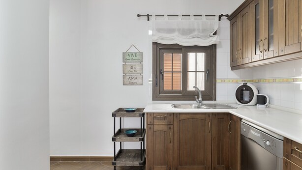 Una cocina con armarios de madera, encimera blanca, un fregadero, una cafetera y un carrito pequeño con cuencos.