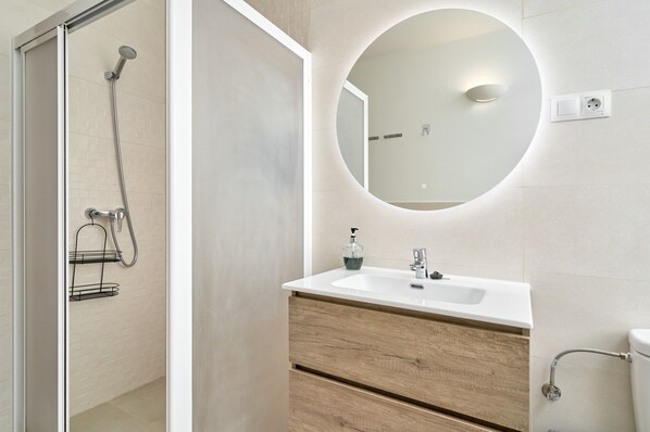 A modern bathroom with a glass shower enclosure, a round mirror with integrated lighting, a white sink with a wooden vanity, and a toilet with a grab bar.
