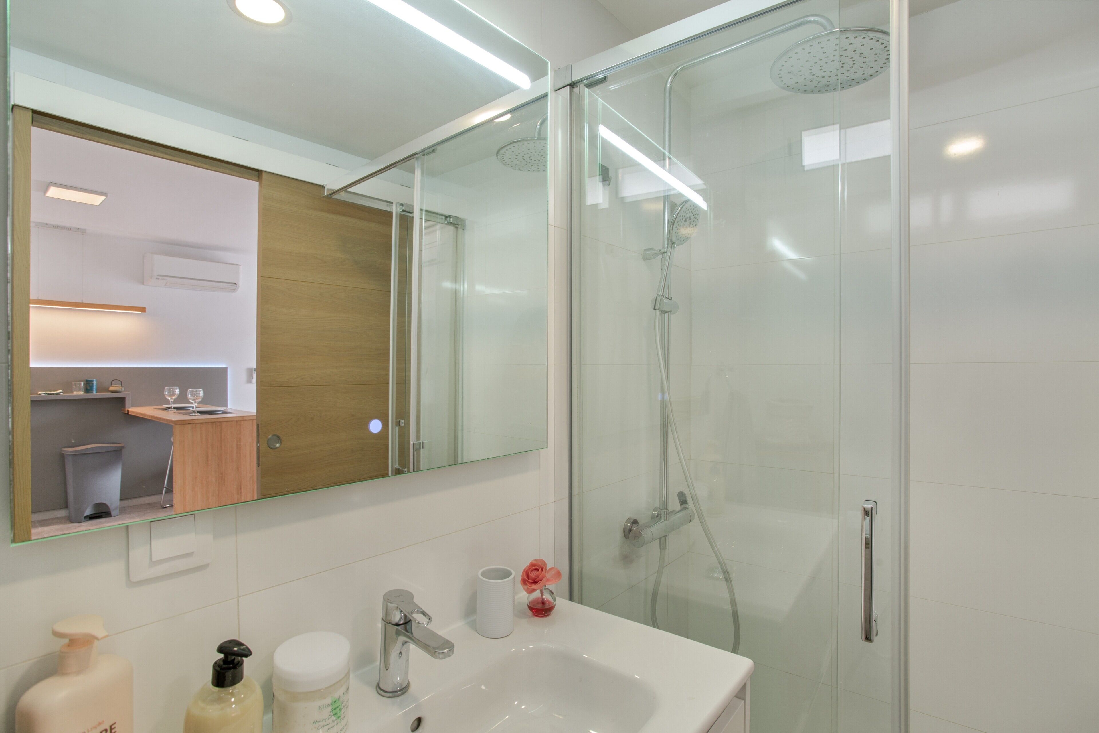 Un baño moderno con cabina de ducha de vidrio, lavabo blanco y un espejo que refleja el ambiente.