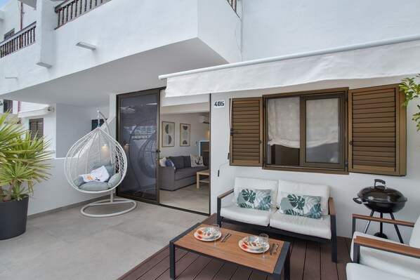 A modern outdoor seating area with a white sofa, a hanging chair, and a wooden table with plates and cutlery.