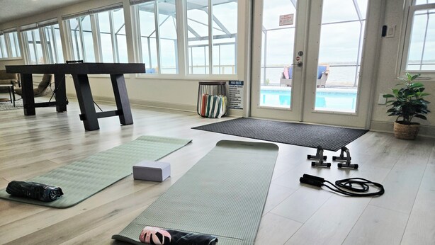 Une pièce dotée de grandes fenêtres, offrant une vue sur la piscine, avec des tapis de sol pour les exercices, une table de massage et une petite plante.