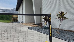 Un terrain délimité par un filet, avec une raquette de pickleball jaune et noire et une balle au sol, devant un bâtiment dont le mur est en briques blanches et le toit est noir.