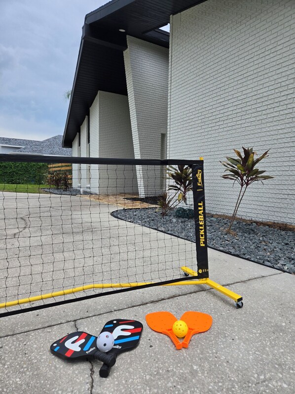 A netted area with a yellow and black pickleball paddle and a ball on the ground, in front of a building with a white brick wall and a black roof.