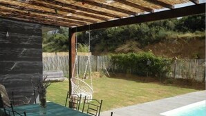 Eine überdachte Terrasse mit Tisch und Stühlen, einem Hängesessel und einem Pool im Hintergrund.