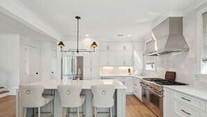 Una cocina moderna con armarios blancos, una isla central con taburetes de bar, electrodomésticos de acero inoxidable e iluminación colgante.